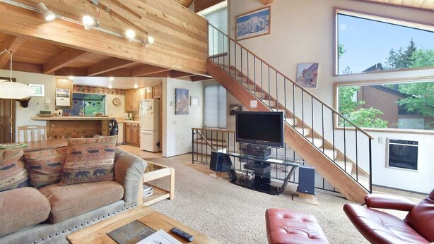 A spacious living area with a high ceiling, wooden beams, a staircase, a television, and a kitchen in the background. - Snowflower 53: Spacious, Partial Mountain Views (Mammoth Lakes)