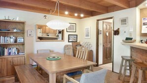 A cozy dining area with a wooden table, benches, and a chair. There is a bookshelf with books and decorative items. A kitchen area is visible in the background with wooden cabinets and a hanging light fixture. - Snowflower 53: Spacious, Partial Mountain Views (Mammoth Lakes)