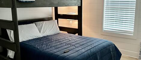 Una cama litera con colcha azul, una mesita de noche con lámpara y una ventana con persianas.