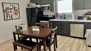 Et moderne køkken med mørke skabe, et rustfrit stål køleskab, en mikrobølgeovn og en opvaskemaskine. Der er et spisebord med stole og en lille plante på bordet.