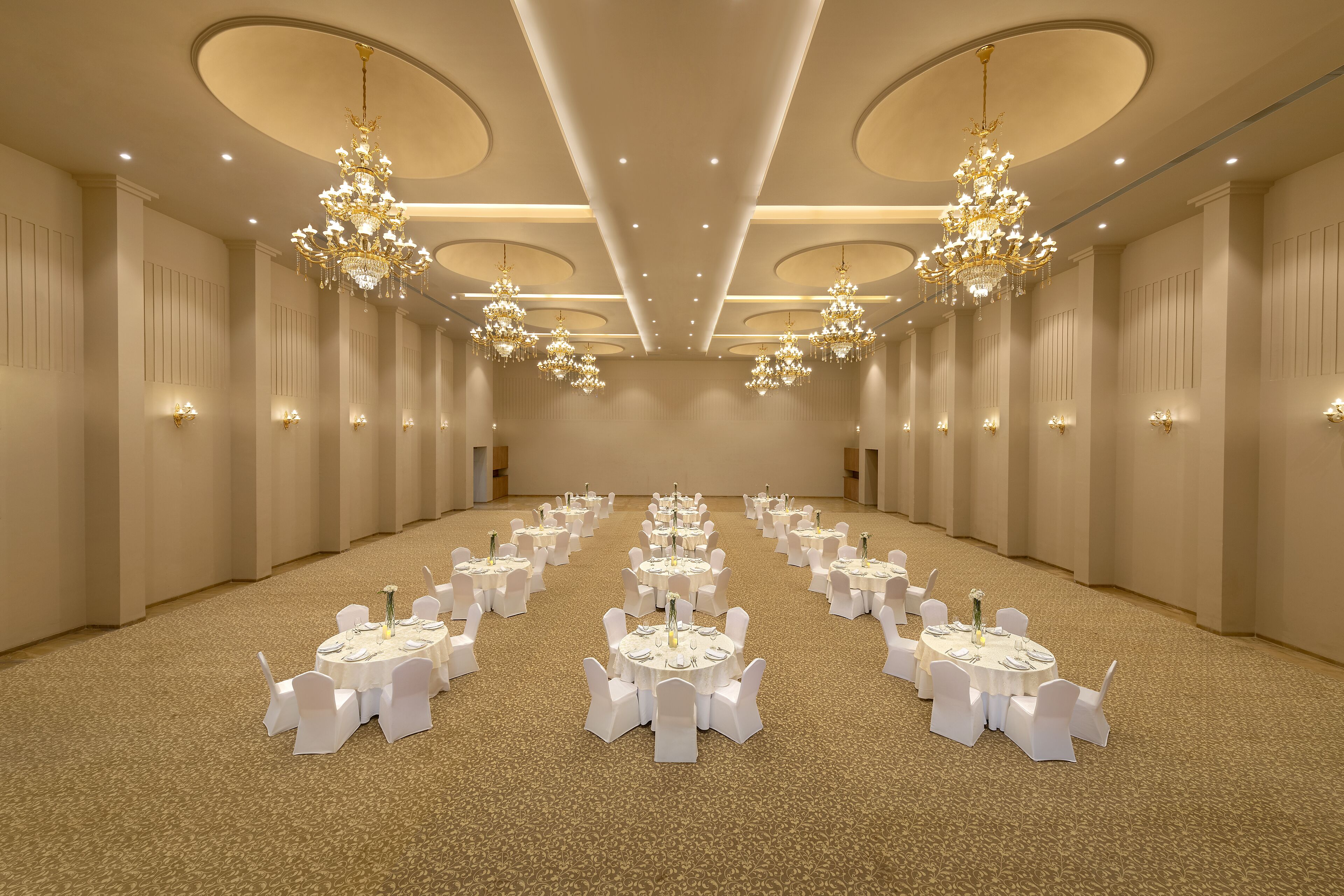 Uma grande sala de banquetes bem iluminada, com várias mesas redondas dispostas para refeições, cada uma rodeada por cadeiras brancas.