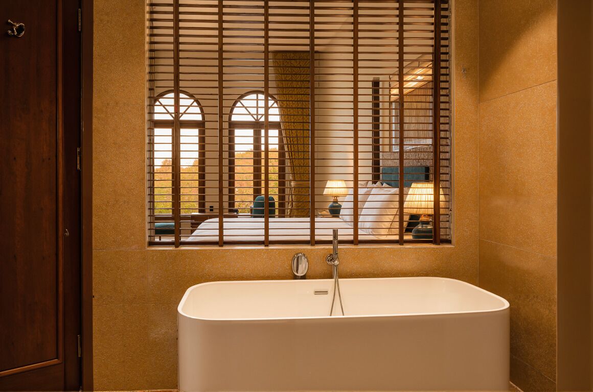 A bathroom with a freestanding bathtub, a window with blinds, and a view of the room.