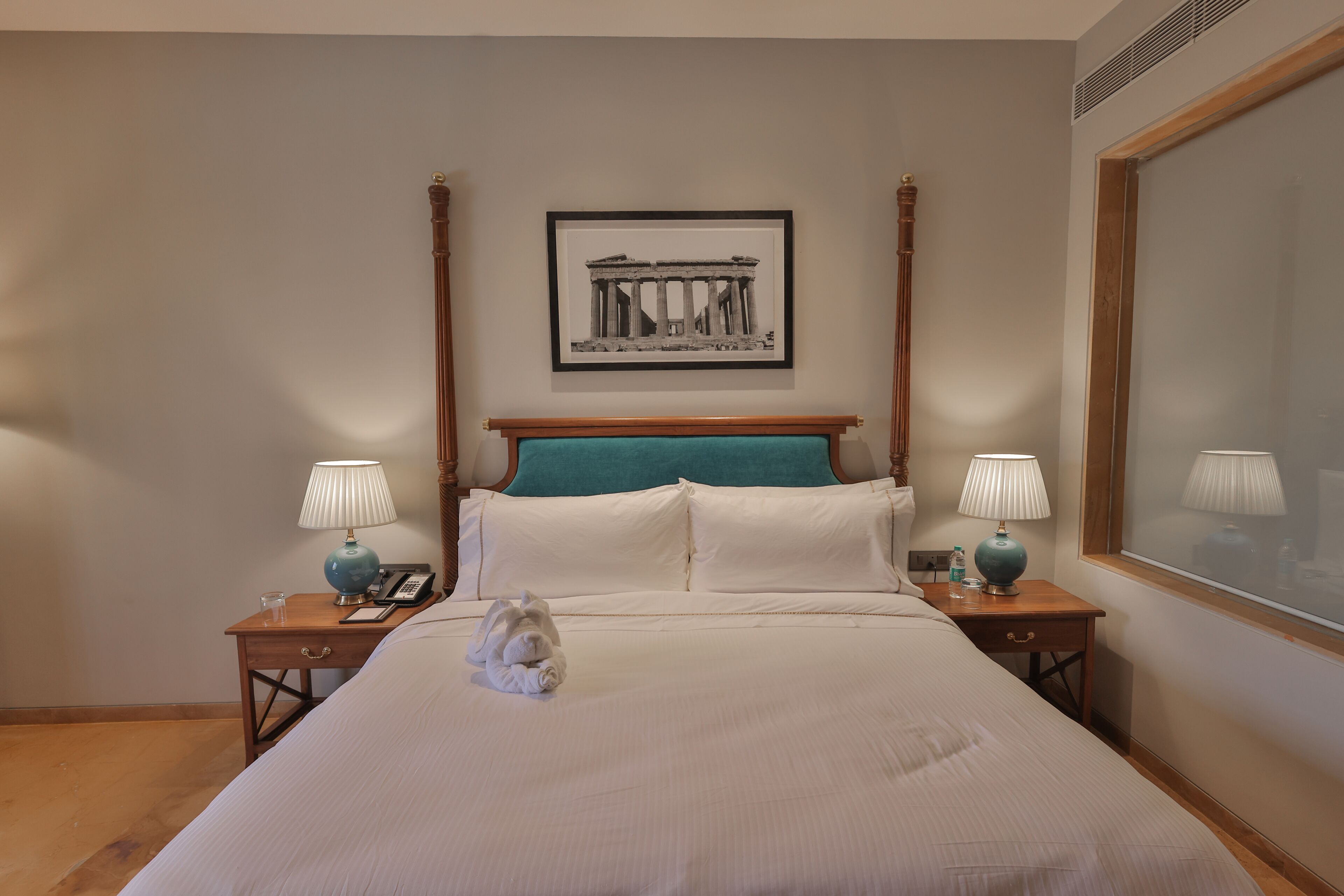 A neatly made bed with white linens, a framed picture of ancient architecture on the wall, and two bedside tables with lamps and a glass of water.