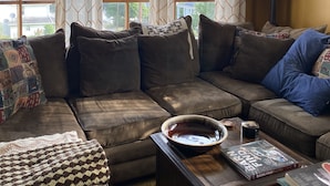 A cozy living room with a large sectional sofa, a coffee table with books and a bowl, and a view of the outdoors through large windows. - Large, Beautiful Mid Century Mountain House..... (South Williamsport)