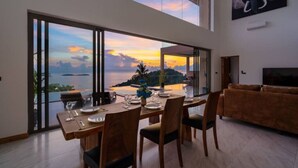 A dining area with a table set for four, large sliding glass doors, and a view of the ocean at sunset. - `Villa Majesta`Amazing View, luxury 4br Pool villa (Koh Samui)