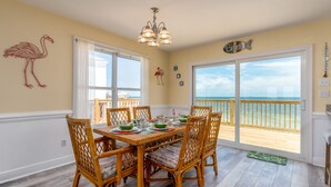 Uma sala de jantar com vista para o oceano, com uma mesa posta para seis pessoas, cadeiras de vime e um lustre.