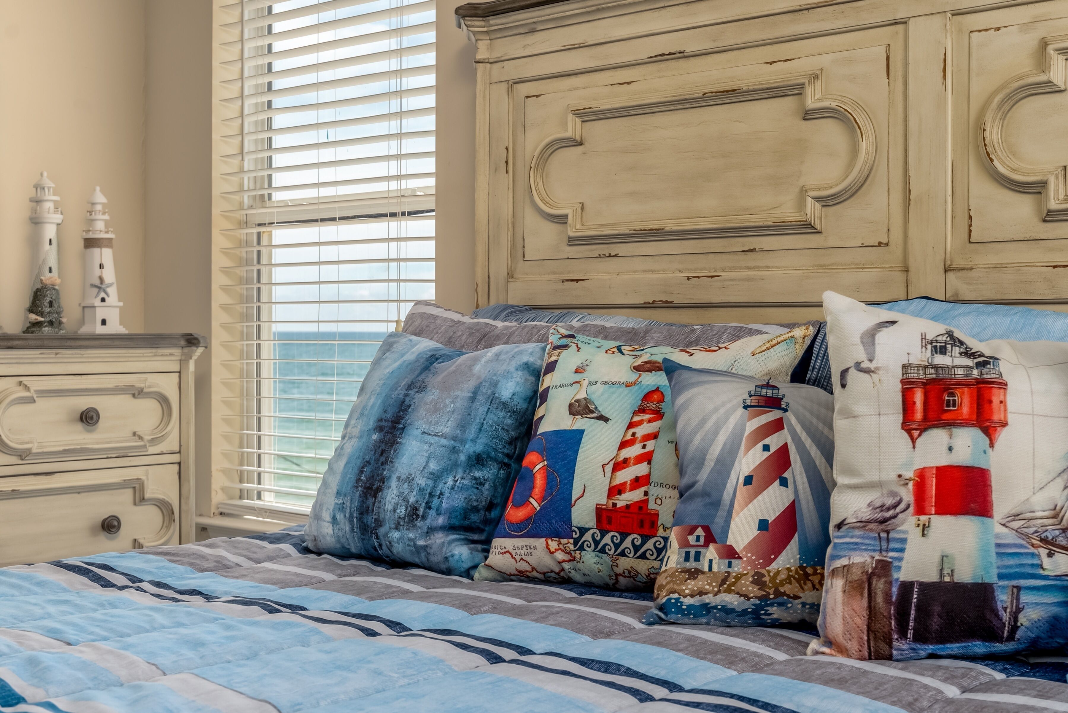 A bedroom with a bed featuring nautical-themed pillows, a dresser with decorative items, and a window with blinds.