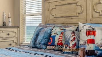 A bedroom with a bed featuring nautical-themed pillows, a dresser with decorative items, and a window with blinds.