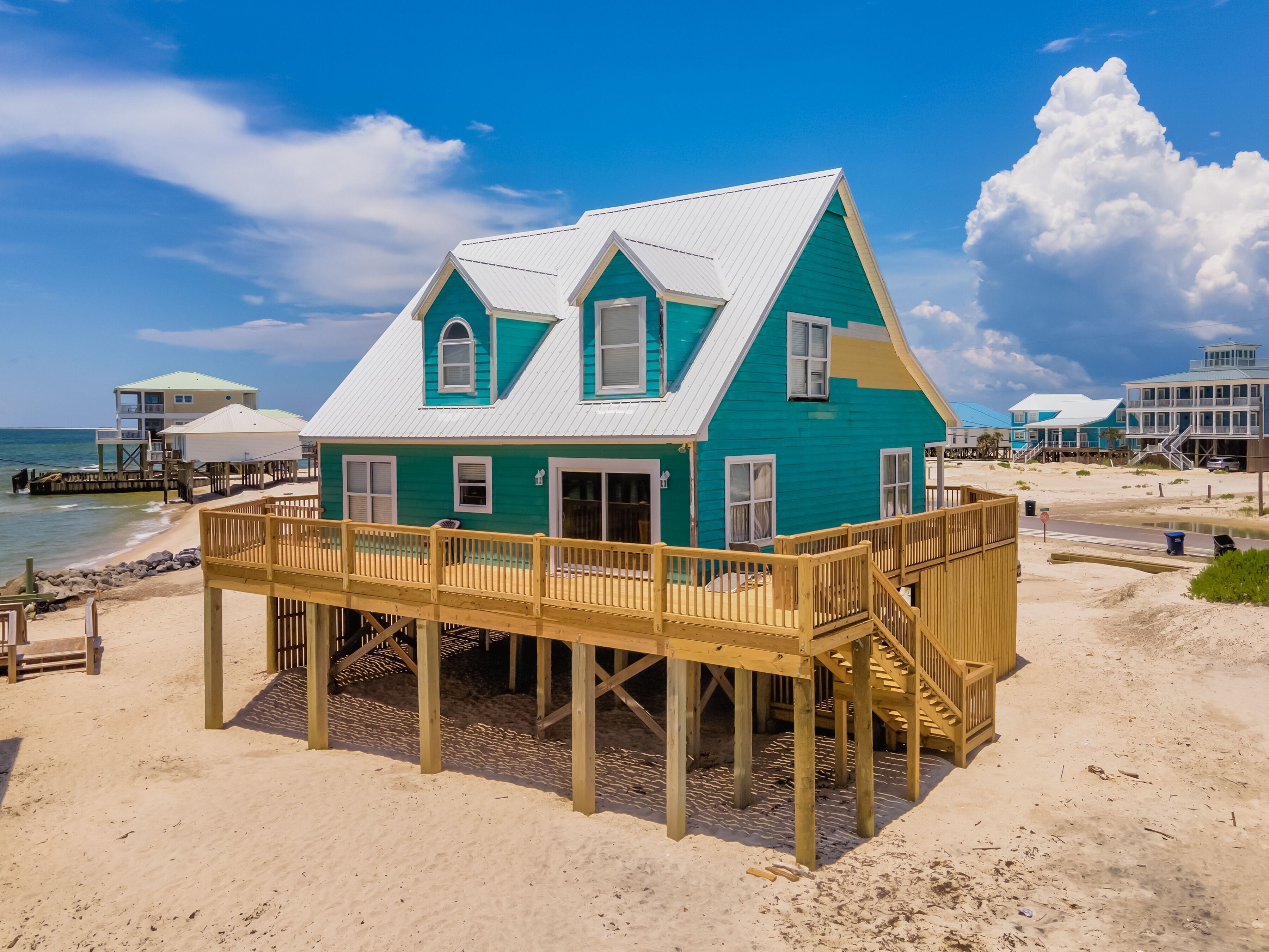 Een turquoise strandhuis met een houten terras en een trap naar de ingang.