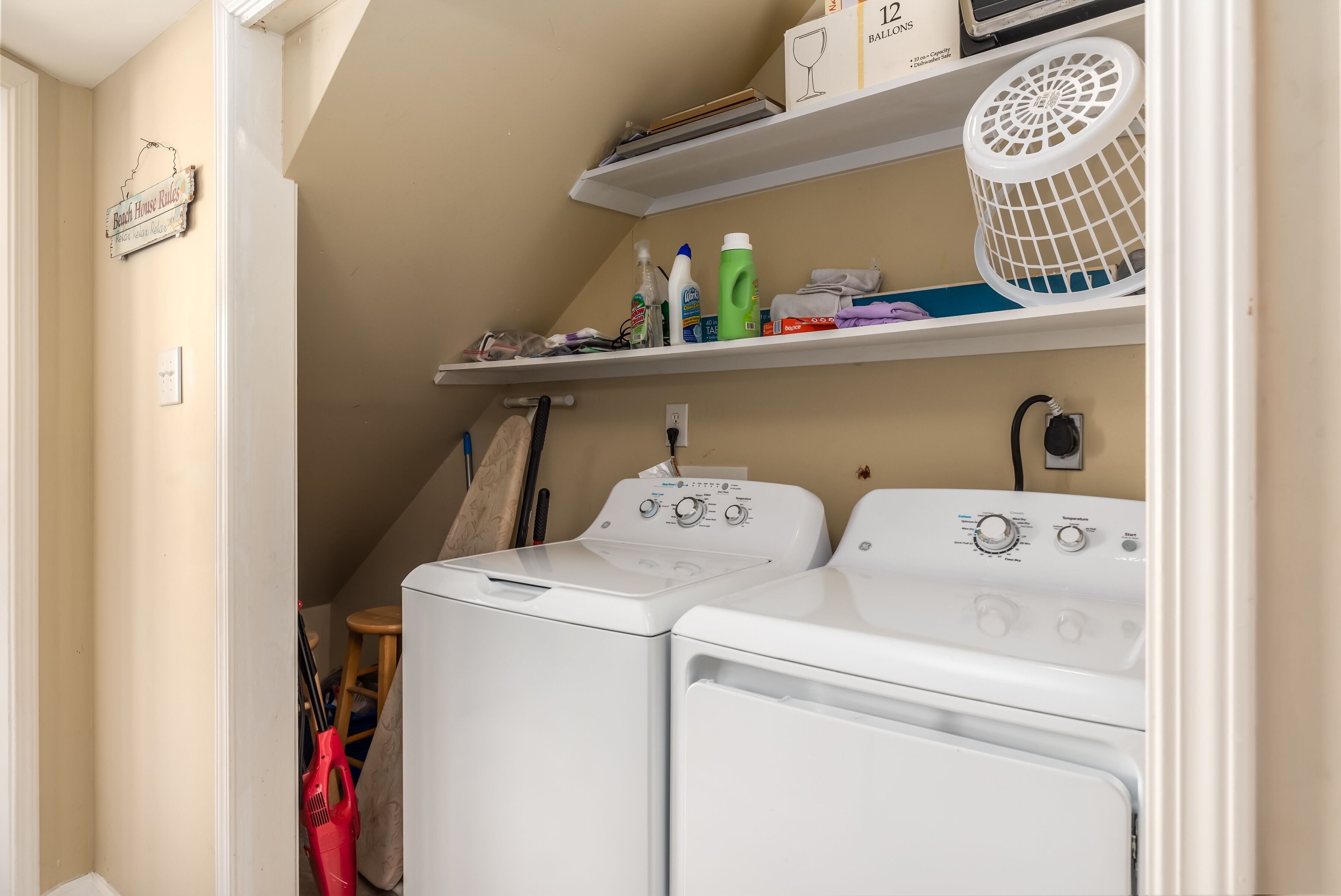 A laundry room with two white front-loading washers and dryers, shelves above holding cleaning supplies, and a sign on the wall.