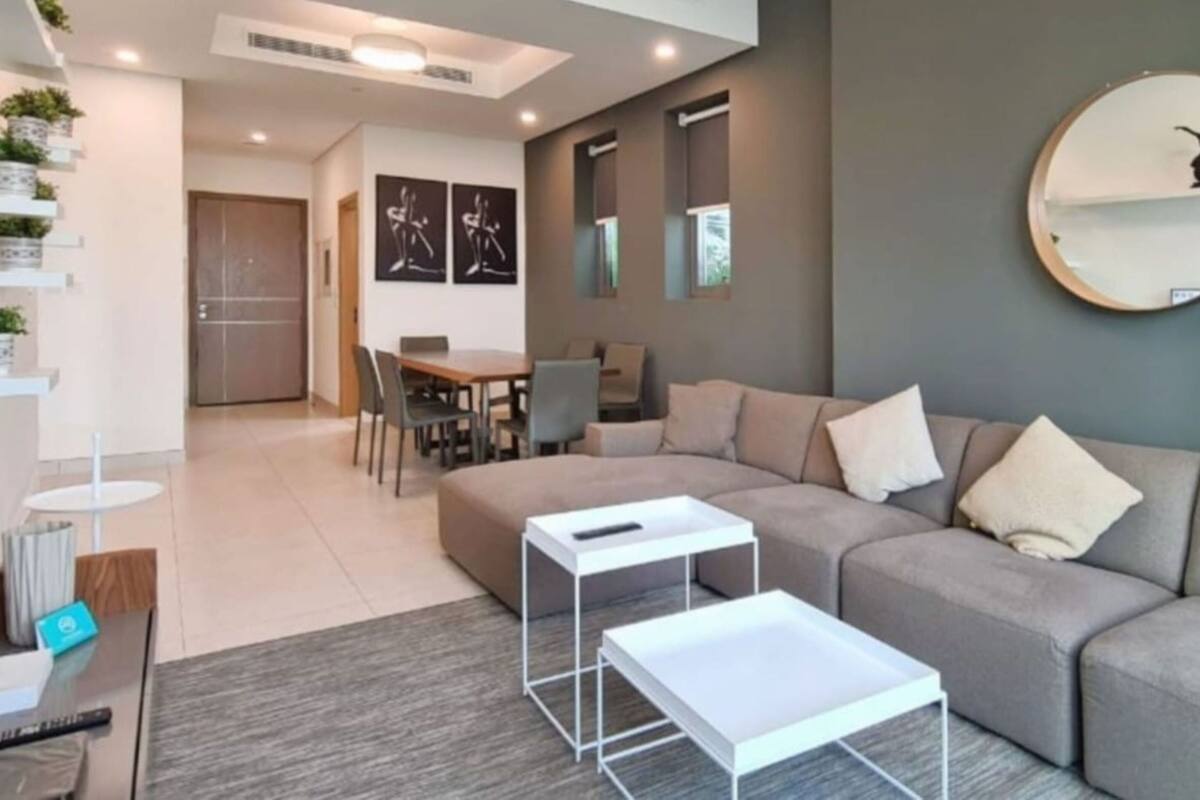 Una sala de estar moderna con un sofá gris, una mesa de centro y un comedor con sillas. Hay estantes empotrados con plantas y obras de arte.