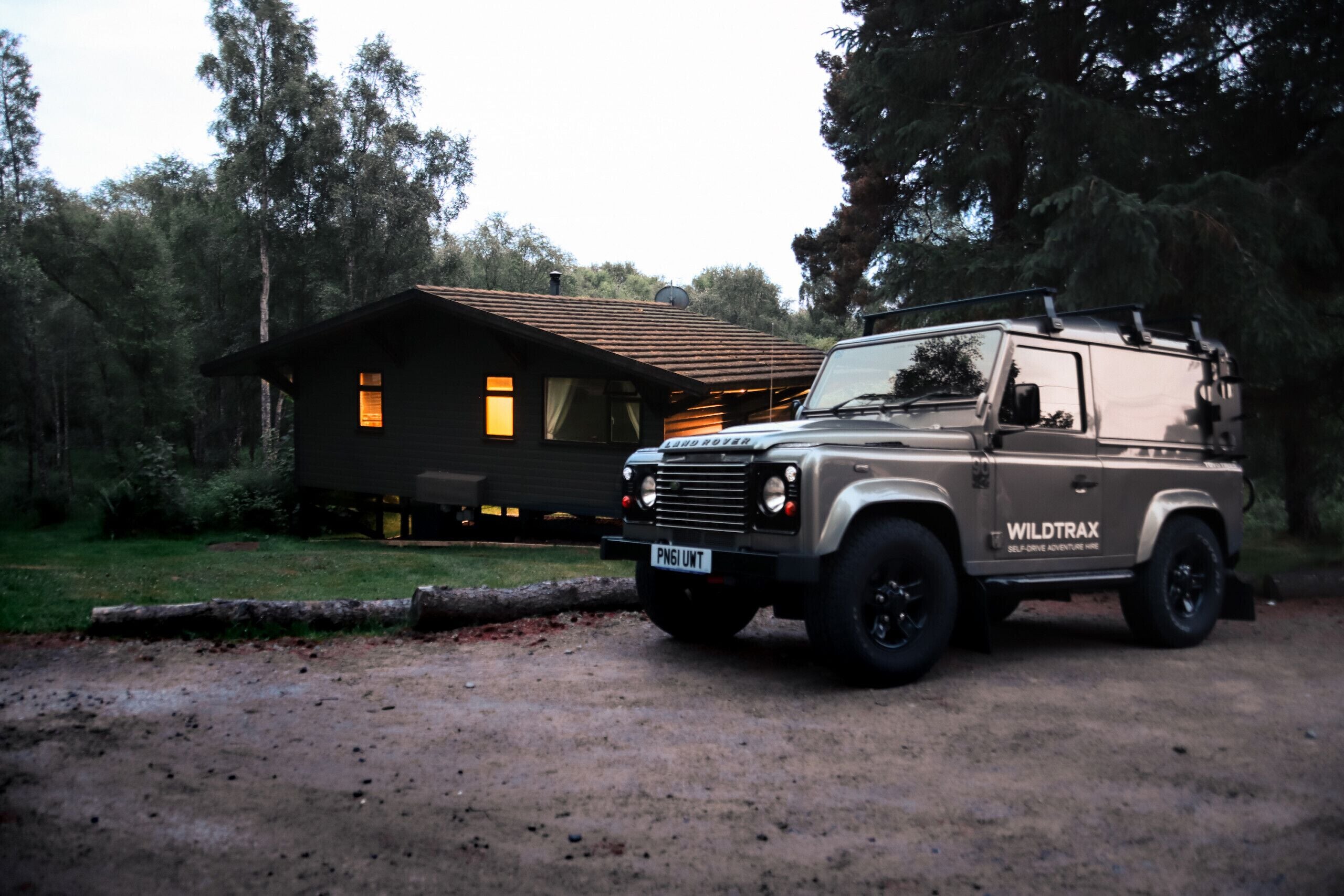 Un vehículo todoterreno color beige con la marca "WILDTRAX" estacionado frente a una cabaña rústica con techo de madera y camino de tierra.