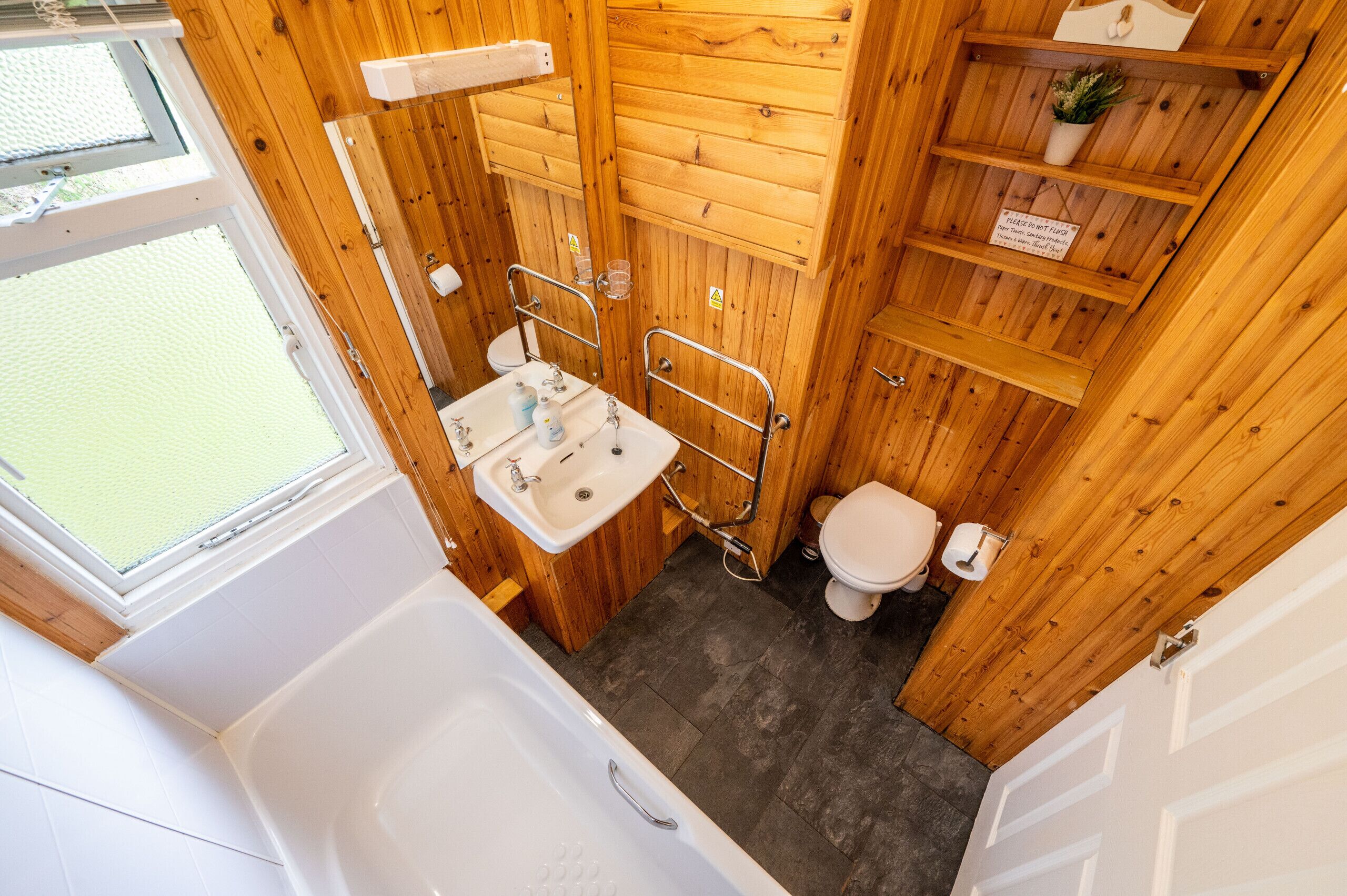 A compact bathroom with a bathtub, sink, and toilet, featuring wooden walls and a small window.
