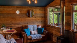 A cozy cabin interior with a wooden wall, a round mirror, a leather sofa, a colorful rug, and a wood-burning stove. - The Wood Hatch Cabin at Ancarraig Lodges, Loch Ness (Drumnadrochit)