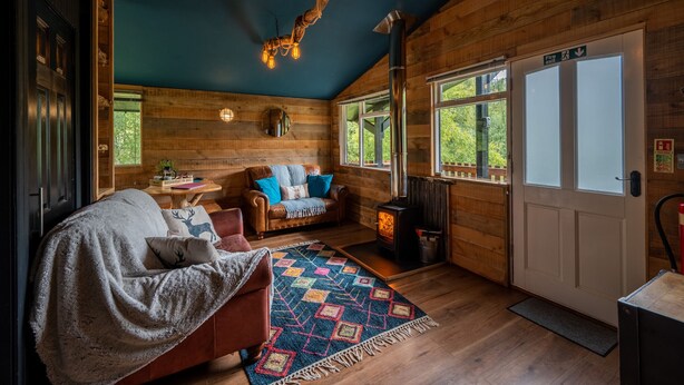 A cozy cabin interior with a fireplace, wooden walls, a sofa, and a dining area. - The Wood Hatch Cabin at Ancarraig Lodges, Loch Ness (Drumnadrochit)