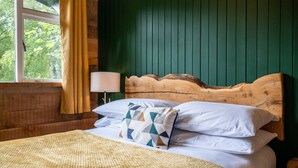 A wooden bed with white bedding and a geometric-patterned pillow, set against a green wall with a window and curtains. - The Wood Hatch Cabin at Ancarraig Lodges, Loch Ness (Drumnadrochit)