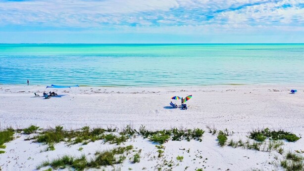 A beach with white sand, clear turquoise water, and sparse vegetation.