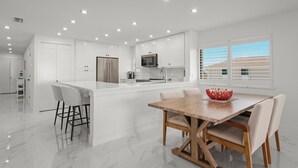 A modern kitchen with white cabinetry, a central island with bar stools, a wooden dining table, and a view of the outside through large windows.