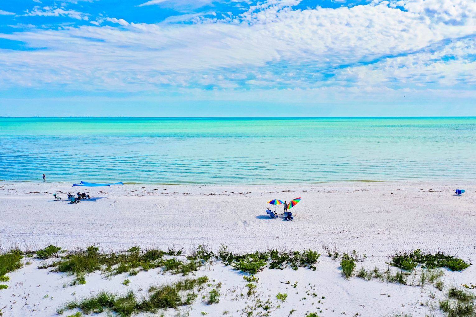 A beach with white sand, clear turquoise water, and sparse vegetation.