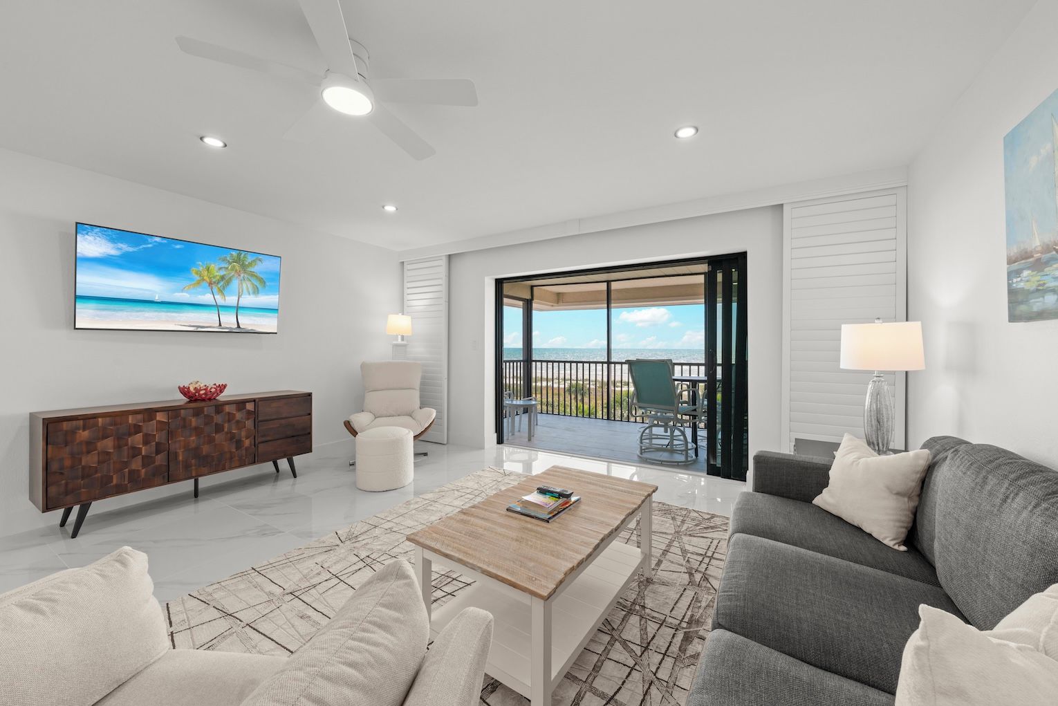 A modern living room with a large TV, a wooden coffee table, a grey sofa, and a balcony with a view of the beach.