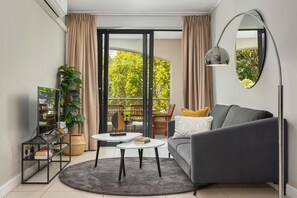A modern living room with a grey sofa, a round coffee table, a TV on a black stand, and a large mirror on the wall. - The Rockwell 104 (Cape Town)