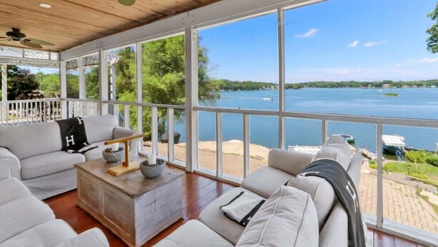 A spacious room with a large window overlooking a lake, furnished with a sofa, a coffee table, and a ceiling fan. - The Mill-Lauderdale Lakes Getaway |Breathtaking Views |Kayaks |15 Minutes to Skiing |Pier |Firepit (Elkhorn)