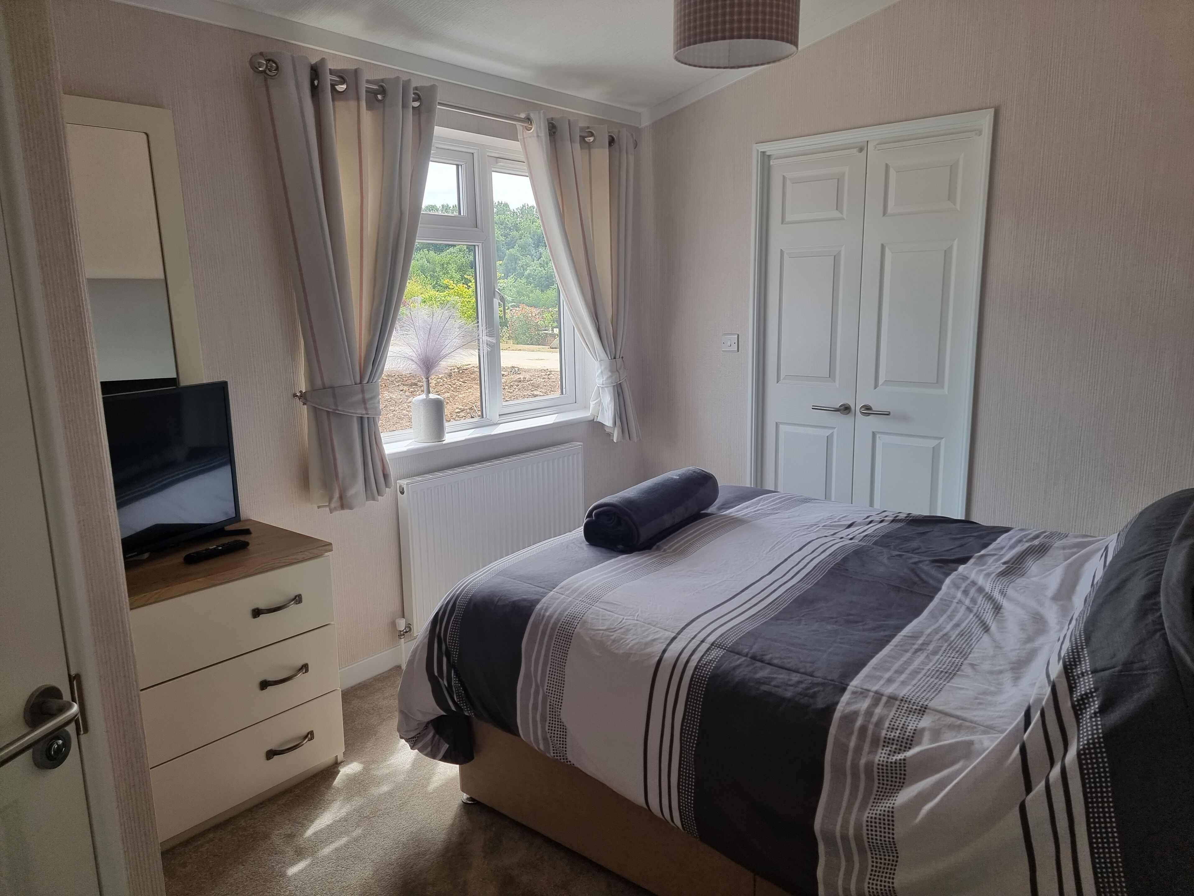 A bedroom with a bed, a TV on a wooden stand, a white door, and a window with curtains.