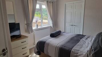 A bedroom with a bed, a TV on a wooden stand, a white door, and a window with curtains.