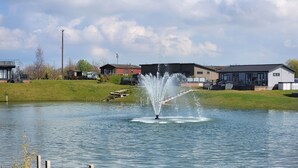 Ein See mit einem Brunnen in der Mitte, umgeben von grünen Wiesen und Gebäuden im Hintergrund.