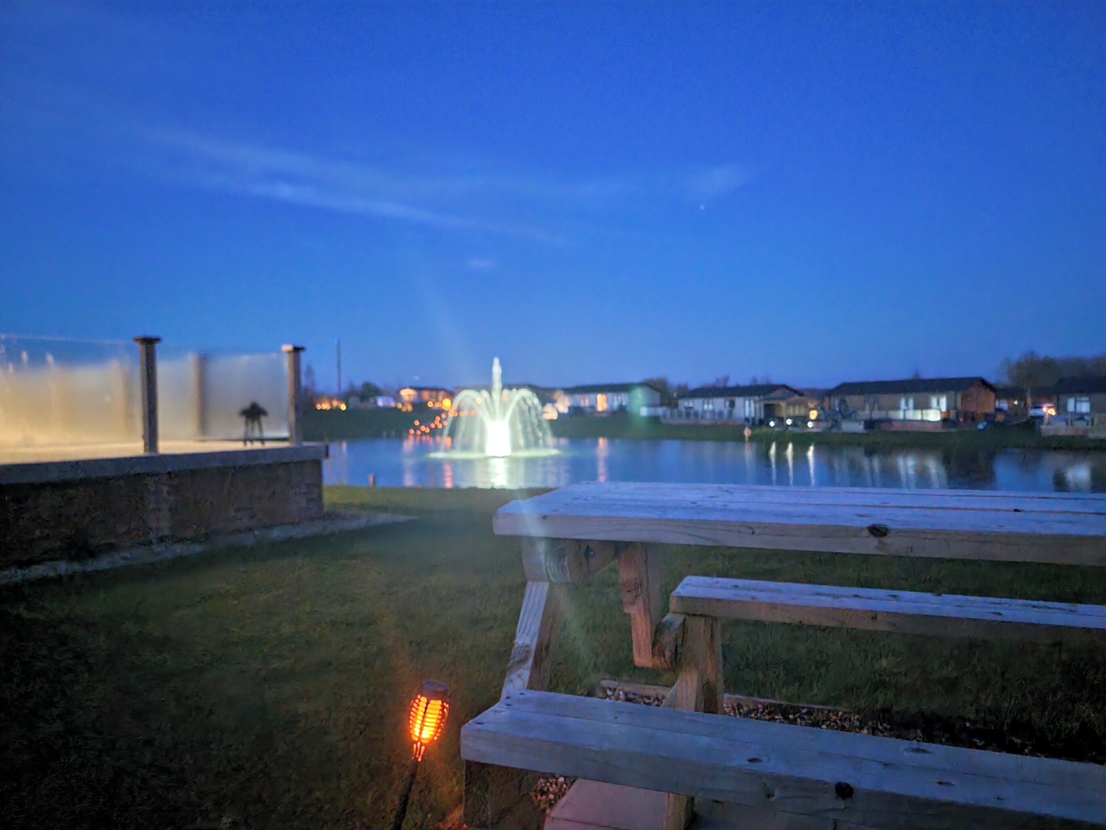 Un parc au bord du lac, doté d’une fontaine, de tables de pique-nique et offrant une vue lointaine sur des maisons.