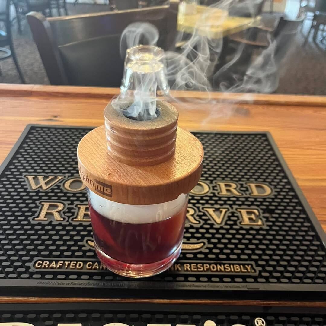 A wooden bottle with a metal cap, filled with a red liquid, placed on a black mat with gold text.
