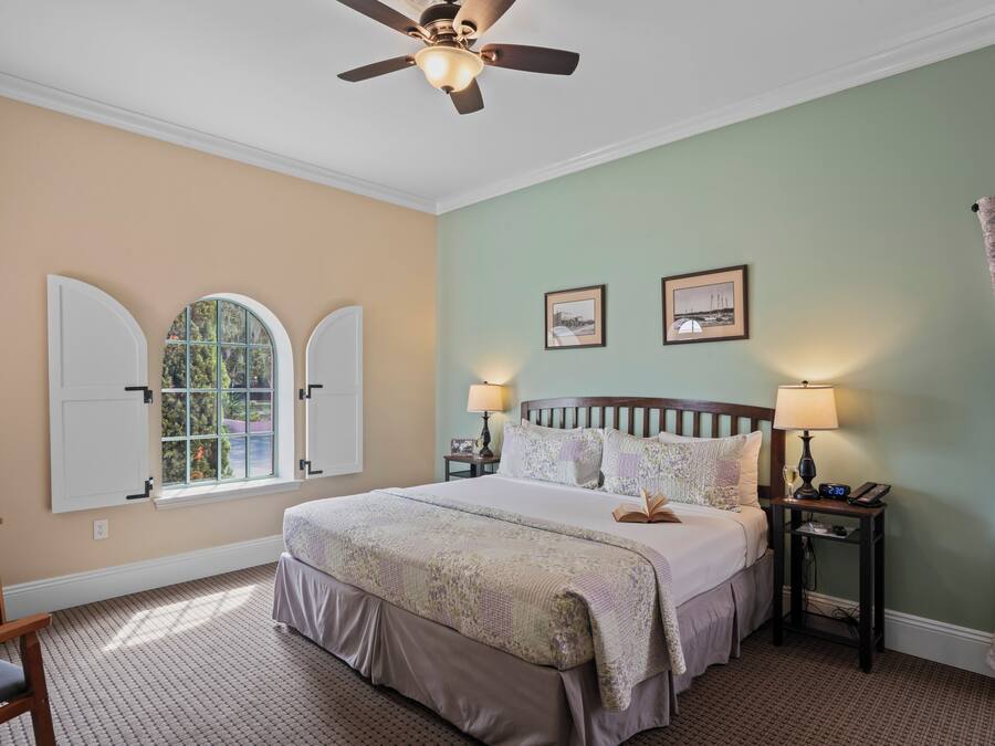 A bedroom with a bed, two bedside tables, a ceiling fan, a window with shutters, and two framed pictures on the wall.
