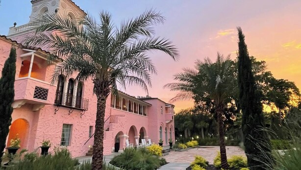 A pink building with arched windows and balconies, surrounded by palm trees and a concrete pathway. - The Hacienda (New Port Richey)