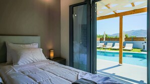 A bedroom with a large bed, a nightstand, and a view of a swimming pool and mountains. - Aurelia Villa (Zakynthos)