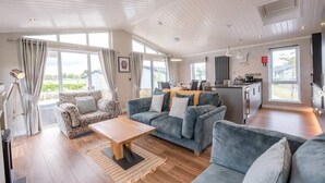 A spacious living area with a wooden coffee table, two grey sofas, and a patterned armchair. A kitchen area is visible in the background with modern appliances. - Marshwiggle, Aldeburgh (Aldeburgh)