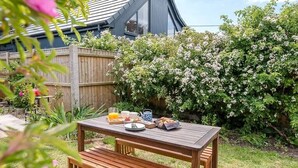 A wooden picnic table with a bench, surrounded by a garden with flowering bushes and a wooden fence.