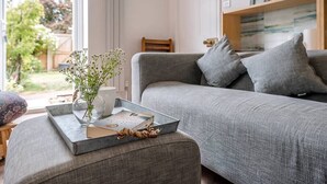 A living room with a grey sofa, a small table with a tray holding a book and a vase with flowers, and a view of the outdoors.