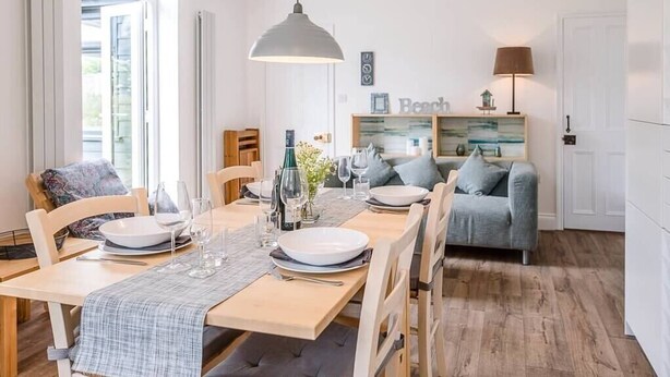 A dining area with a wooden table set for four, a grey sofa, and a wall-mounted shelf with decorative items.