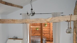 Una habitación con techo de madera, una cama con manta gris, un sofá azul con cojines, un tocador blanco y una mesita con un libro.