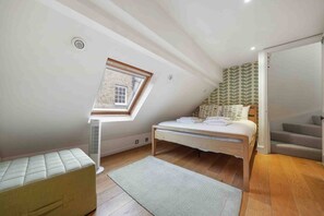 Une chambre avec un plafond en pente, un puits de lumière, un lit avec des oreillers, une table de chevet, une lampe sur pied et un escalier.