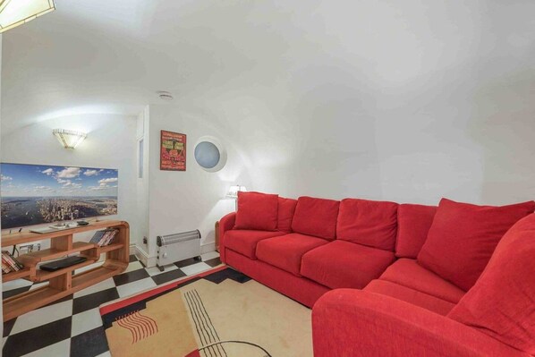 Un salon avec un canapé rouge, une bibliothèque en bois, un tapis à carreaux et une affiche représentant un paysage urbain accrochée au mur.