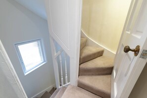 A staircase with beige carpeted steps leading to a landing, a white door with a brass doorknob, and a window with blinds on the left wall.