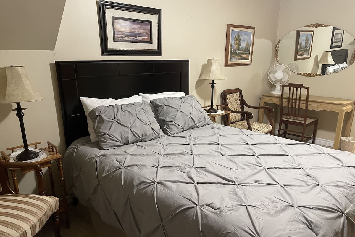 A neatly made bed with a grey quilt, a bedside table with a lamp, a chair, and a mirror on the wall.