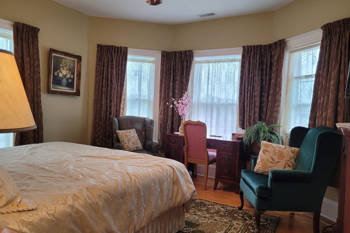 A bedroom with a large bed, two chairs, a desk, and a window with curtains.