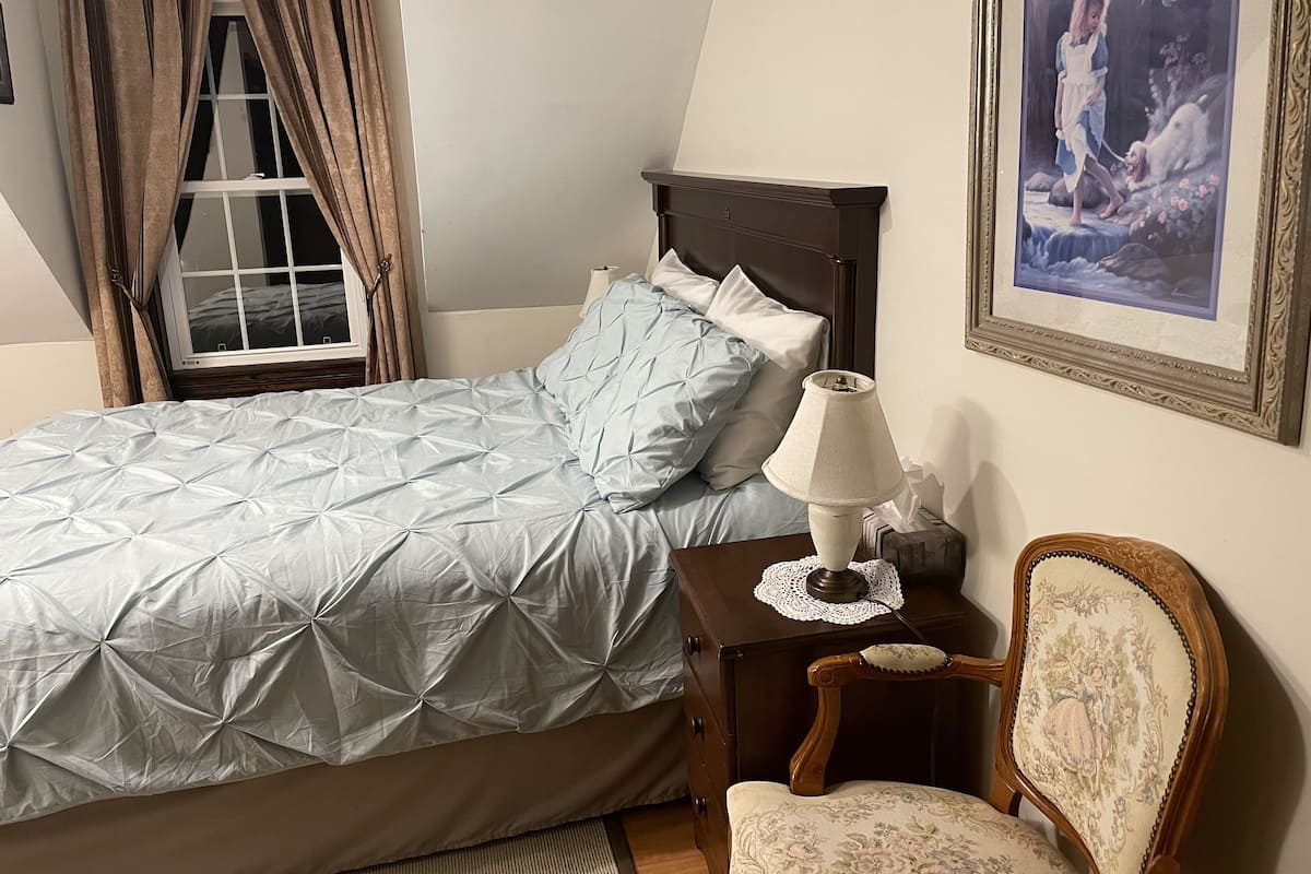 A neatly made bed with a blue quilt, a bedside table with a lamp, a chair with floral upholstery, and a framed picture on the wall.