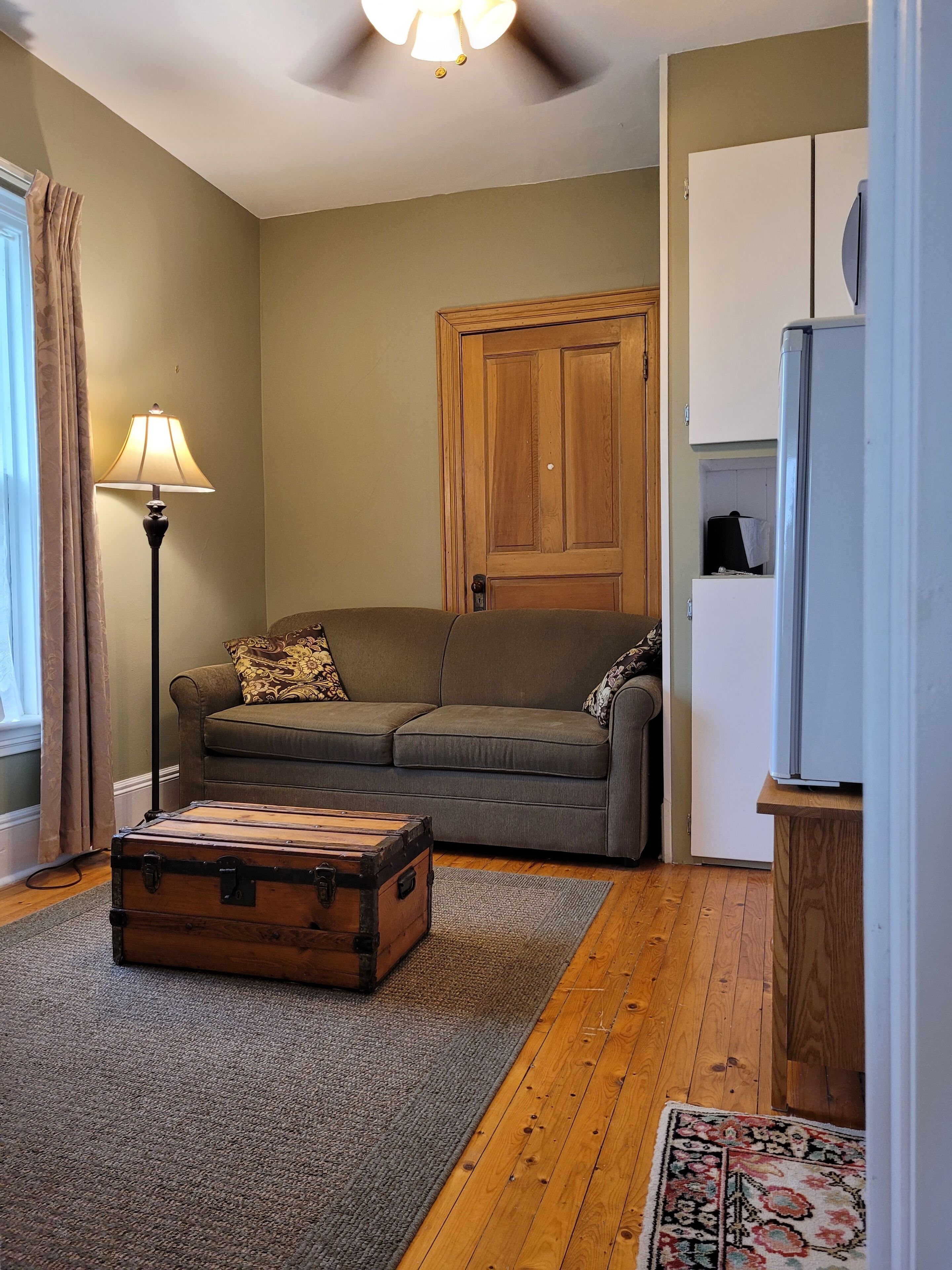 Una sala de estar con piso de madera, un sofá verde, una lámpara, una puerta de madera y un refrigerador.