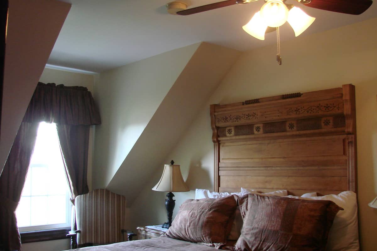 A bedroom with a sloped ceiling, a bed with multiple pillows, a nightstand with a lamp, and a window with curtains.