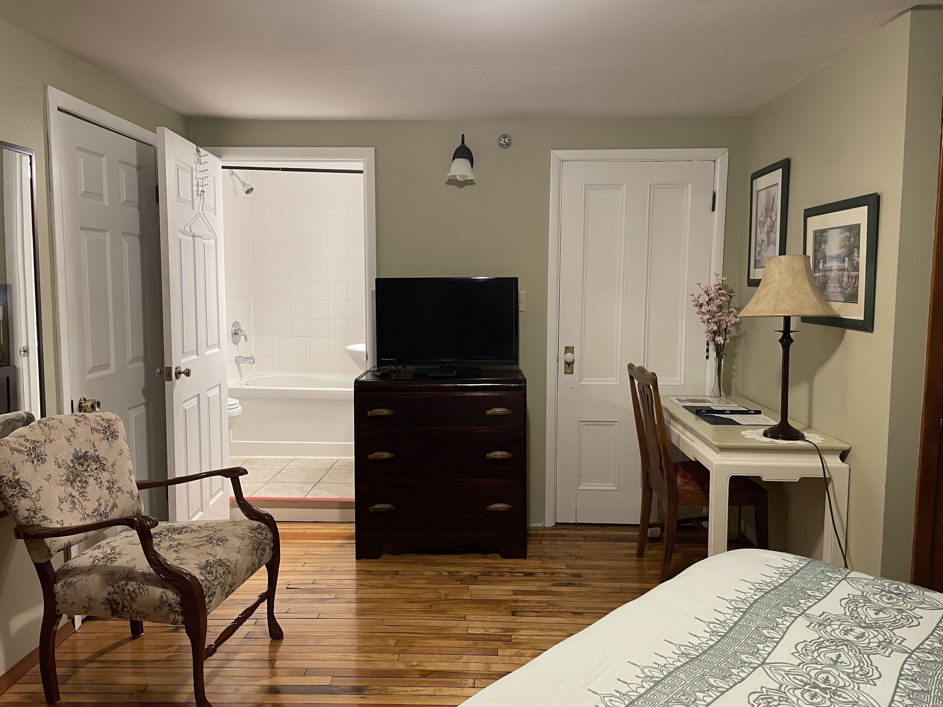 A bedroom with a bed, a dresser, a chair, a desk with a lamp, a television, and a door leading to a bathroom.