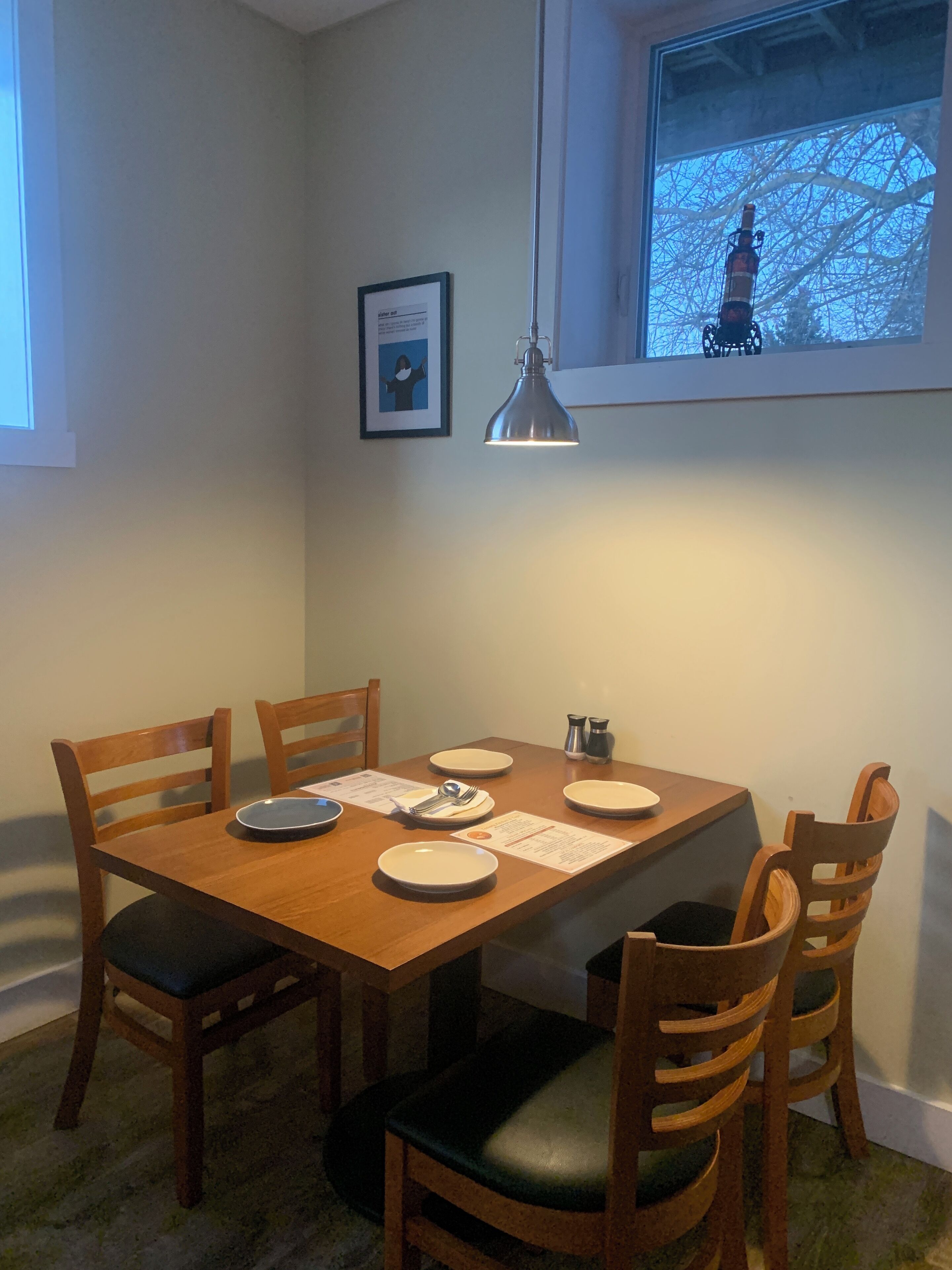 A dining area with a wooden table, four chairs, and a pendant light. There is a window with a view of a tree and a statue.