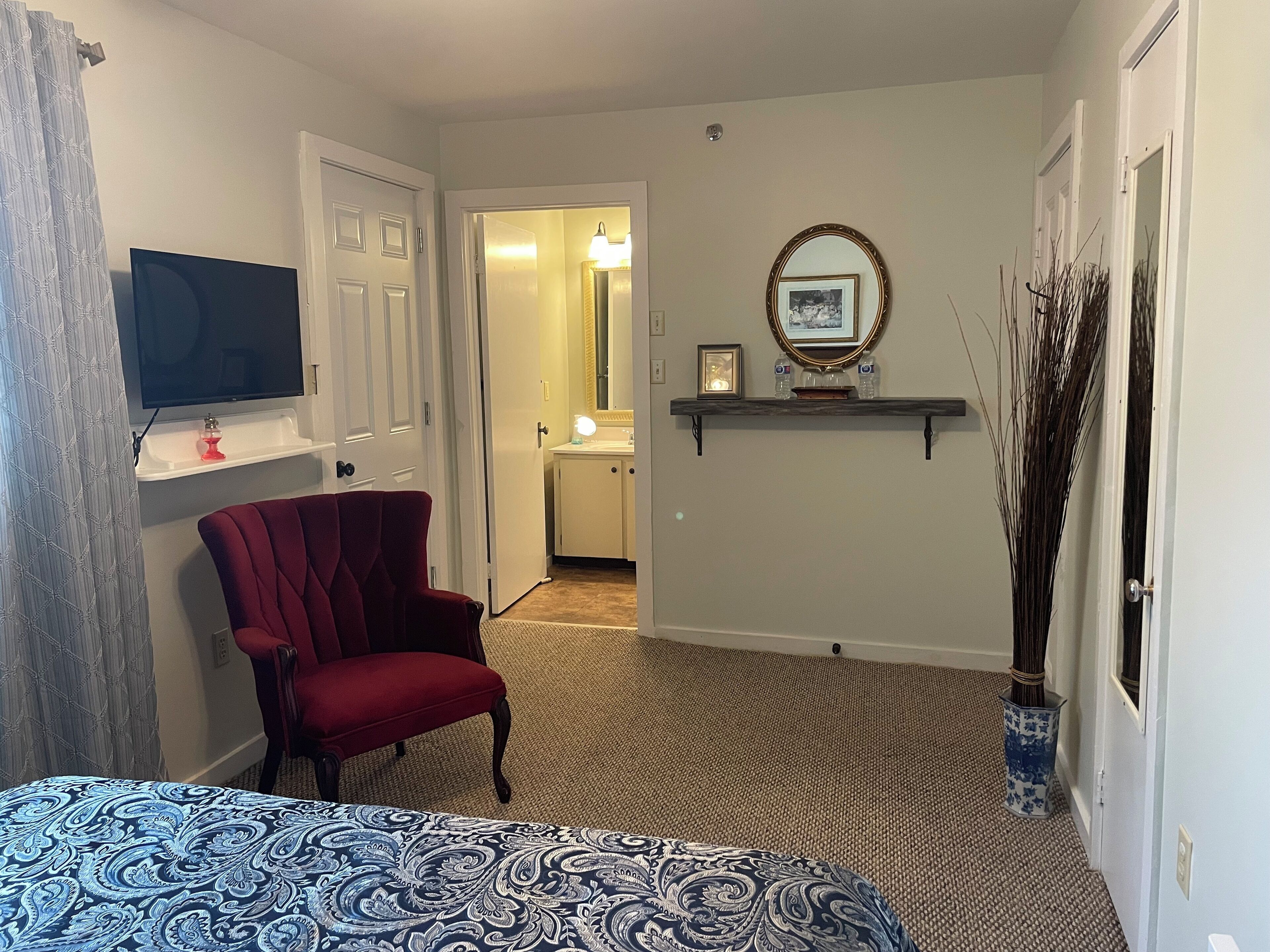 Habitación de hotel con un sillón rojo, una cama con colcha estampada, un televisor, un espejo y un pasillo que conduce a un baño.